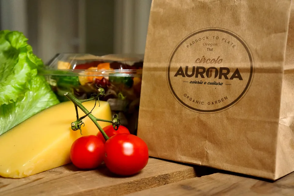 Un sacchetto di carta kraft con il marchio Circolo Aurora stampato a timbro, accanto a insalata, formaggio e pomodori