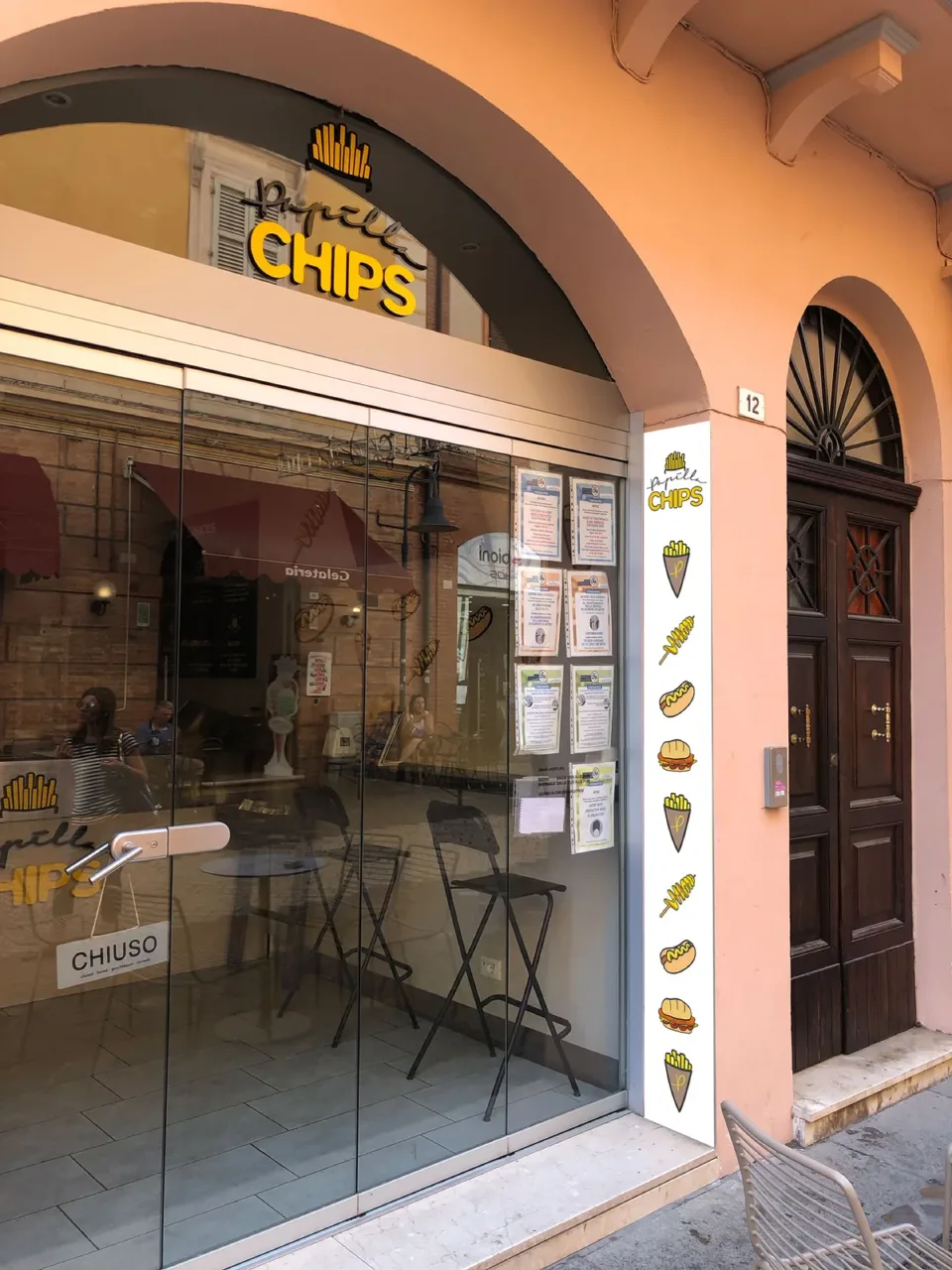 L’ingresso del negozio sotto il portico: l’insegna Papilla Chips sopra la vetrina e, accanto alla porta, il pannello verticale bianco con il marchio e le icone dei prodotti