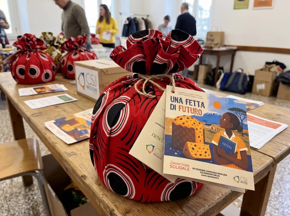 Il panettone solidale nella confezione di tessuto africano rosso e nero, legata con lo spago, con il pieghevole Una fetta di futuro appeso; dietro, sul tavolo, altri panettoni e i volontari