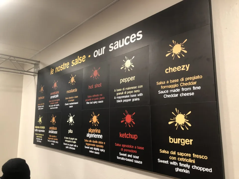 Il pannello nero delle salse sulla parete del negozio, con il titolo le nostre salse – our sauces e dodici riquadri con una macchia di colore e la descrizione in italiano e inglese