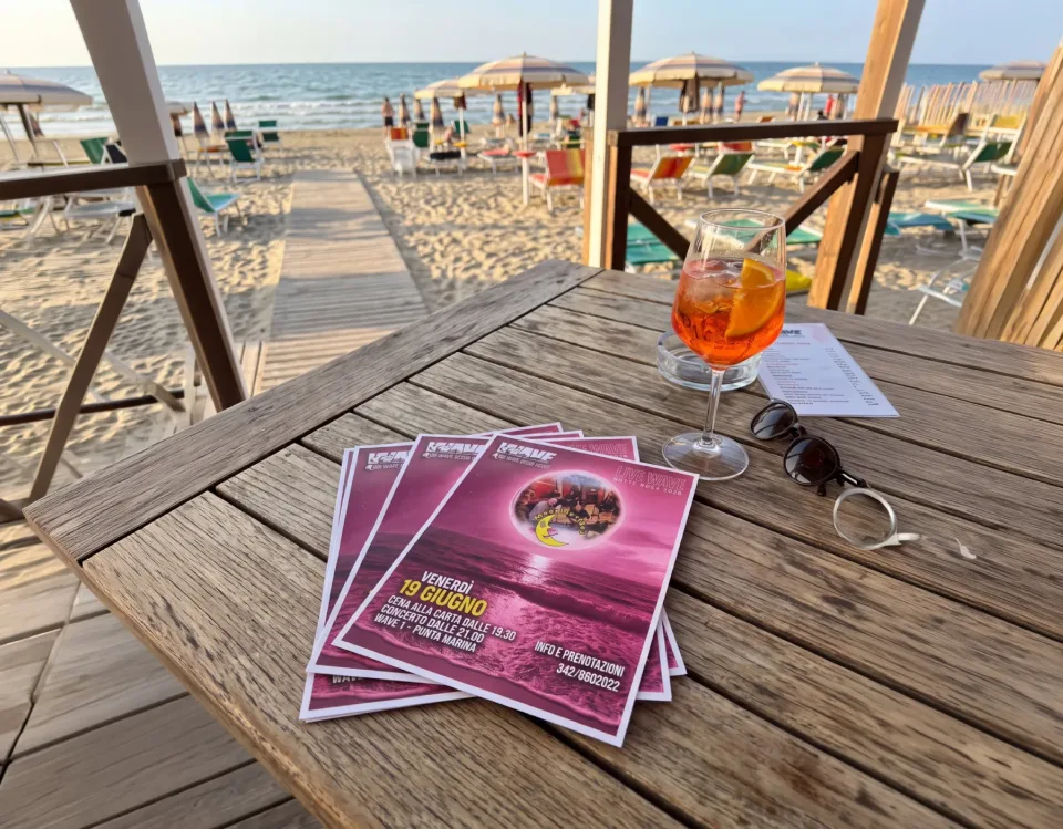 Volantini rosa della serata Live Wave su un tavolo di legno, accanto a uno spritz, con la spiaggia del Wave sullo sfondo