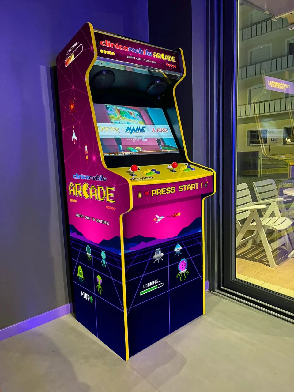 Il cabinet Clinica Mobile Arcade in una stanza dalle pareti viola, accanto a una vetrata, con lo schermo acceso