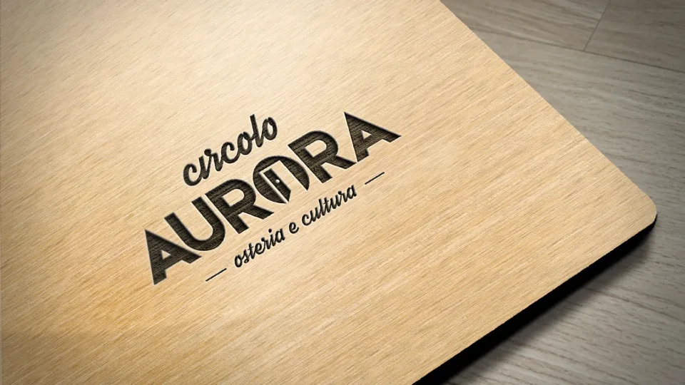 Il marchio Circolo Aurora inciso su una tavola di legno chiaro: la parola circolo in corsivo, AURORA in maiuscolo con una porta rossa socchiusa al posto della O, e la scritta osteria e cultura
