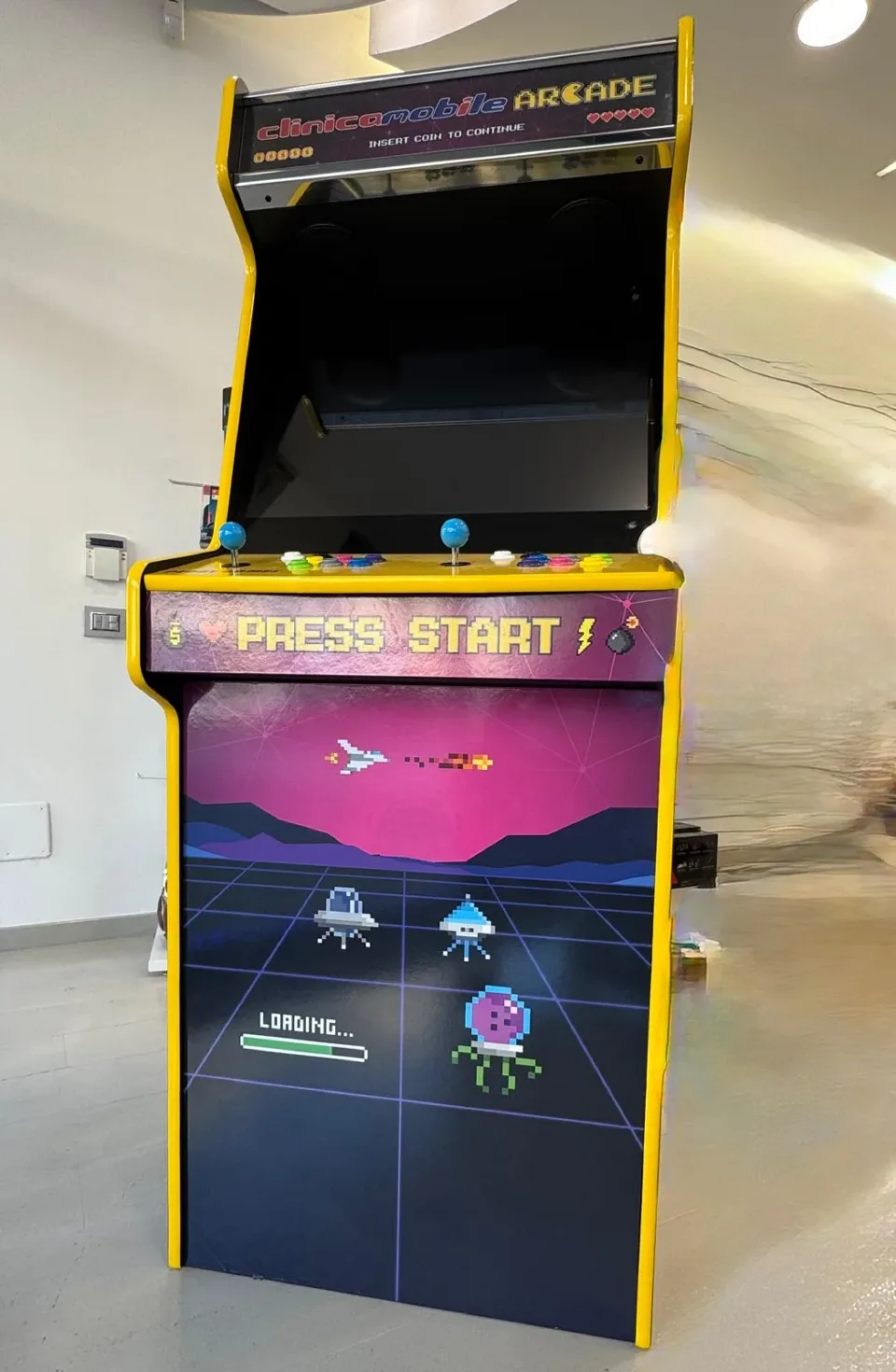 Il cabinet visto di fronte: Press start sul pannello e, nel frontale, una griglia al neon con astronavi in pixel art