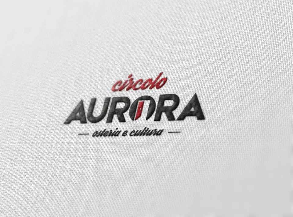 Il marchio Circolo Aurora in rilievo su un tessuto bianco: circolo in rosso, AURORA in grigio scuro con la porta rossa nella O